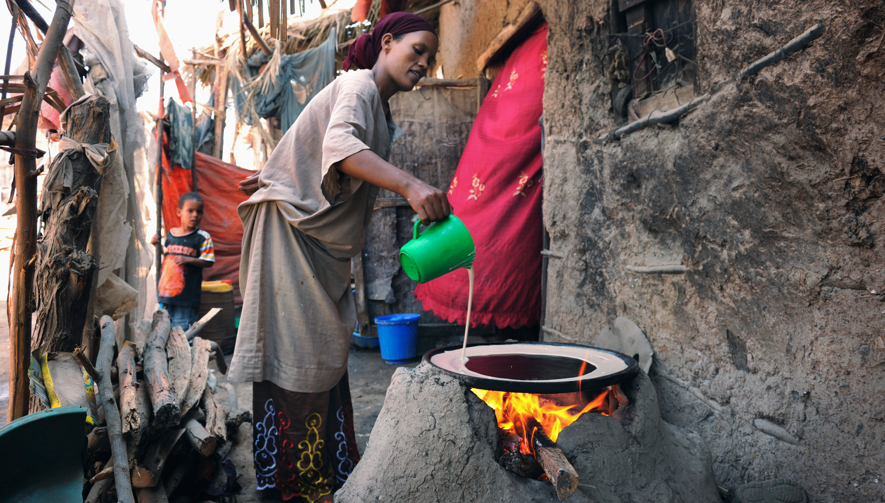 Color photo shows a woman using a biomass-burning oven to make bread outside of a rough-hewn house in Ethiopia