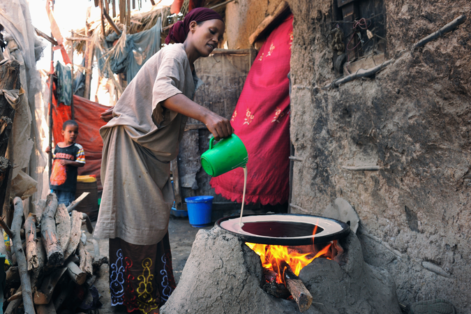 Color photo shows a woman using a biomass-burning oven to make bread outside of a rough-hewn house in Ethiopia