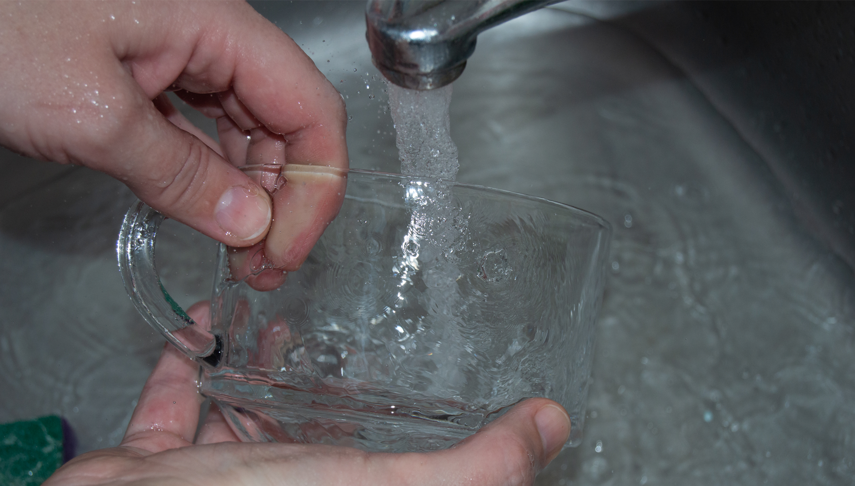 Photo of a person's hands washing a glass mug under the faucet in a stainless-steel sink, with water from the tap forming viscous patterns over all the objects