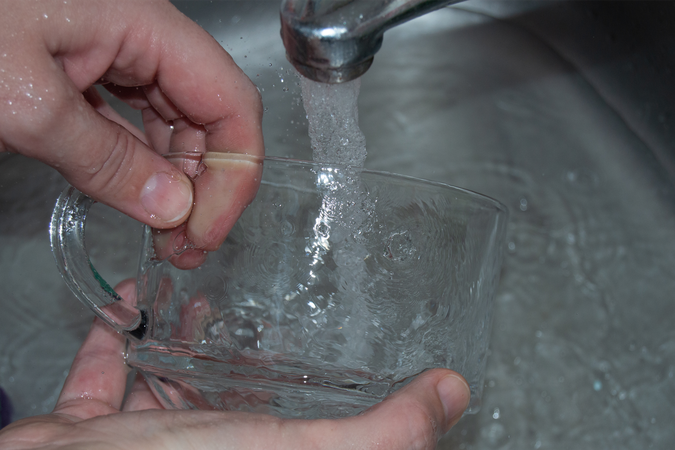 Photo of a person's hands washing a glass mug under the faucet in a stainless-steel sink, with water from the tap forming viscous patterns over all the objects