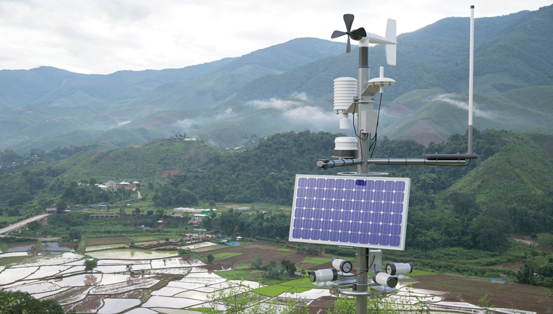 Color photo of a weather station in a rice field, weather station in the foreground and agricultural fields and cloudy mountains in the background