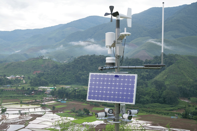 Color photo of a weather station in a rice field, weather station in the foreground and agricultural fields and cloudy mountains in the background