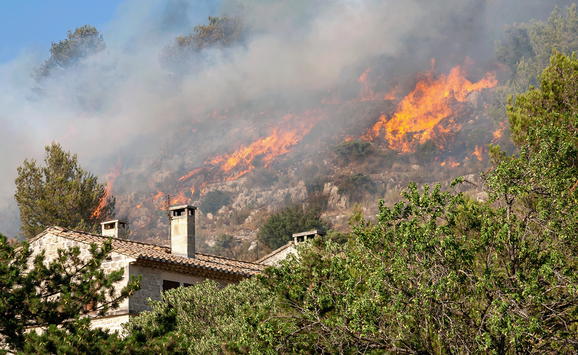 A photo of an untouched house and trees, while flames and smoke from a forest fire approach from the background.