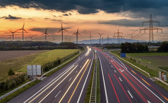 Long-exposure image of cars driving on highway with farms, powerlines, and wind turbines in the background