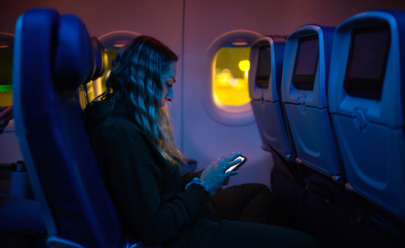 Young women sits in a commercial airplane at night while looking at her bright phone screen.