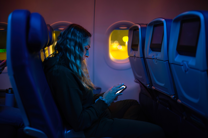 Young women sits in a commercial airplane at night while looking at her bright phone screen.