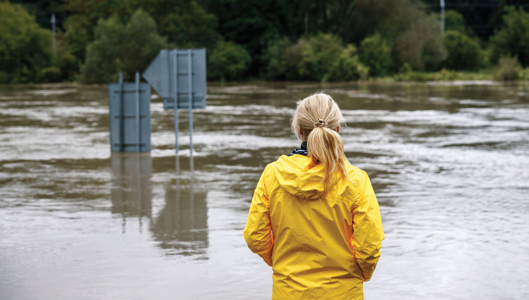 Color photo of a worried woman in a raincoat looking out at overflowing water during a flood, with street signs submerged and peeking up over the water's surface