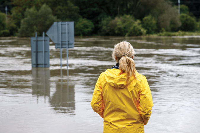 Color photo of a worried woman in a raincoat looking out at overflowing water during a flood, with street signs submerged and peeking up over the water's surface