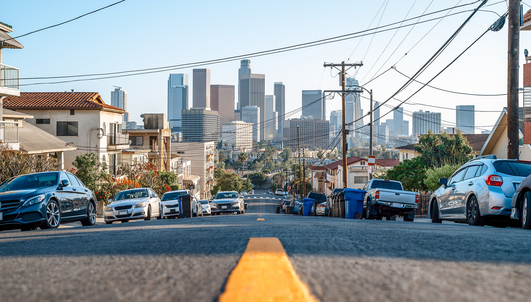 Cars lined up on the side of a street with the skyline of Los Angeles in the background on a clear day