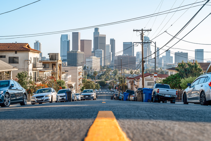 Cars lined up on the side of a street with the skyline of Los Angeles in the background on a clear day