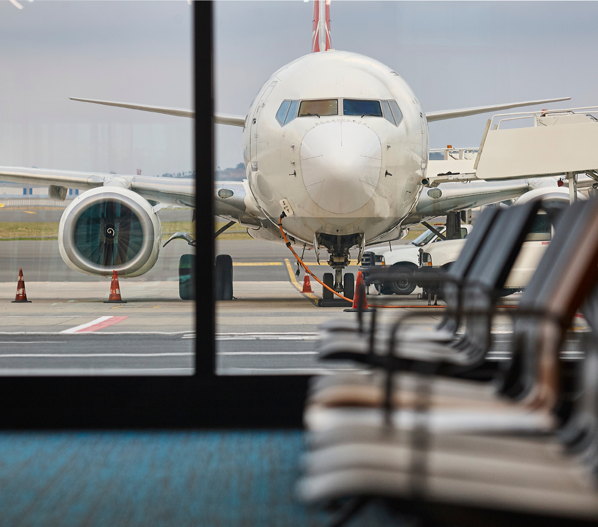 An airplane is visible refueling through the windows of an airport terminal, with empty chairs in the foreground