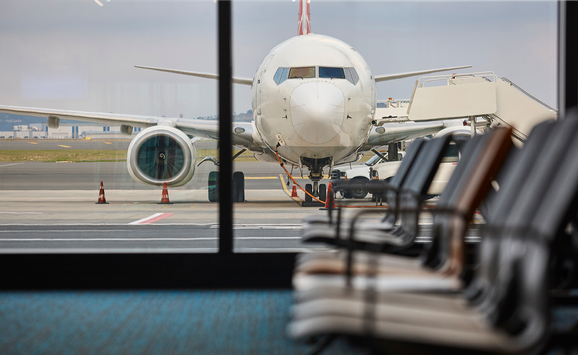 An airplane is visible refueling through the windows of an airport terminal, with empty chairs in the foreground