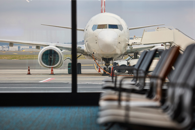 An airplane is visible refueling through the windows of an airport terminal, with empty chairs in the foreground