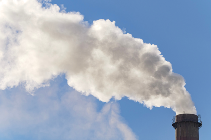 A stream of gray smoke billowing from thermal power plant chimneys, creating a haze against a blue sky.