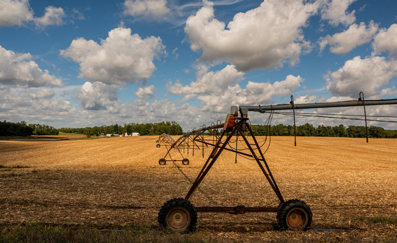 A photo of a harvested corn field, with an irrigation machine in the foreground and farm buildings in the background.