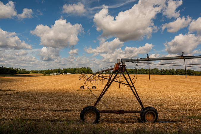 A photo of a harvested corn field, with an irrigation machine in the foreground and farm buildings in the background.