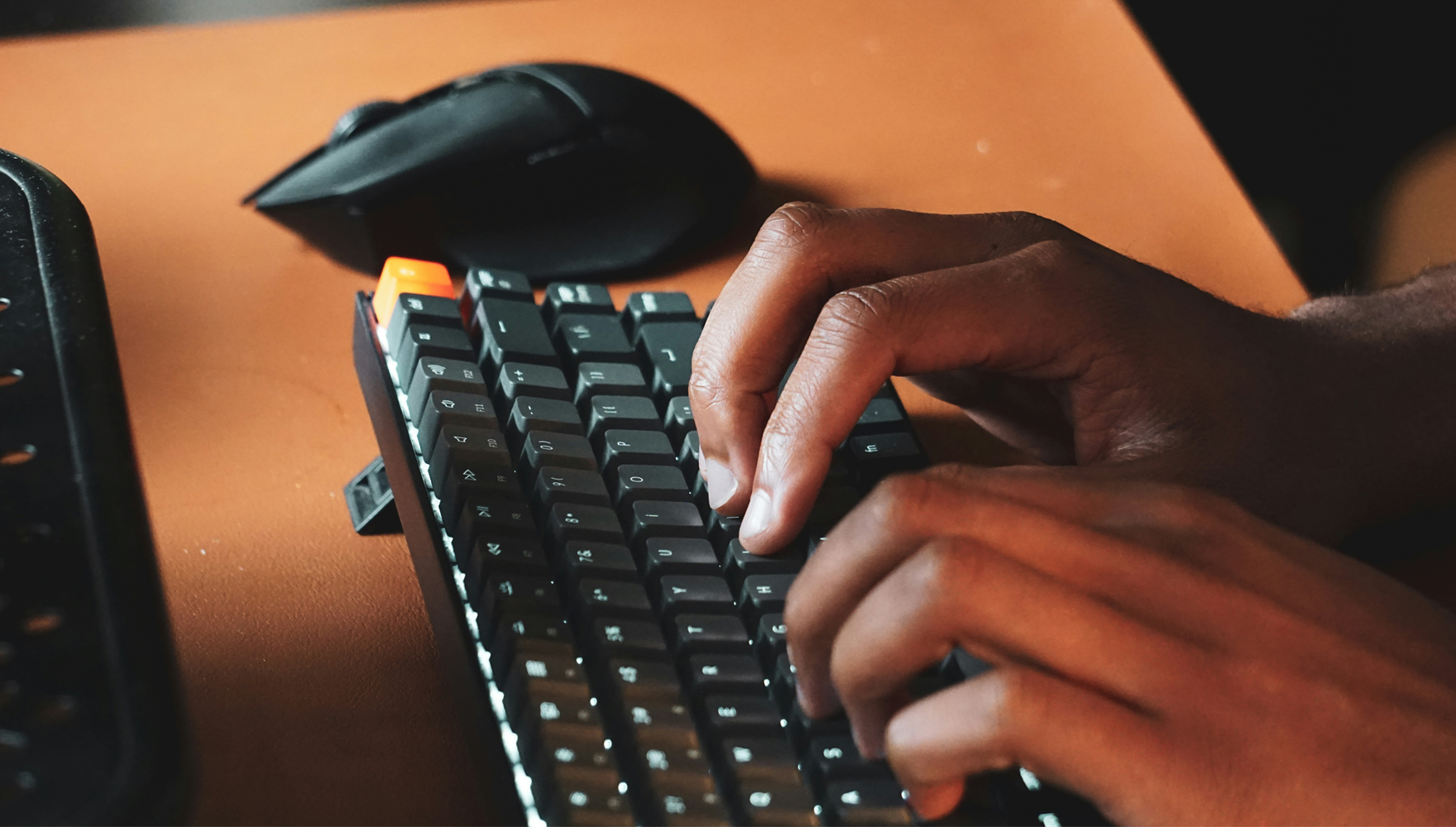 Closeup photo of a person of color typing on a keyboard, with a computer mouse in the background