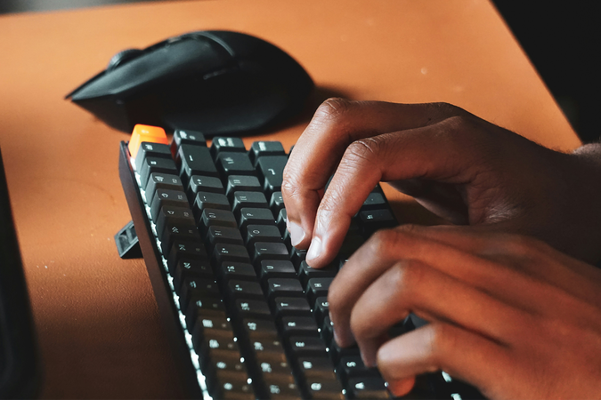 Closeup photo of a person of color typing on a keyboard, with a computer mouse in the background